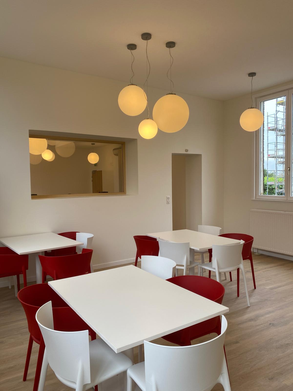 Salle à manger rénovée avec tables blanches, chaises rouges et suspensions